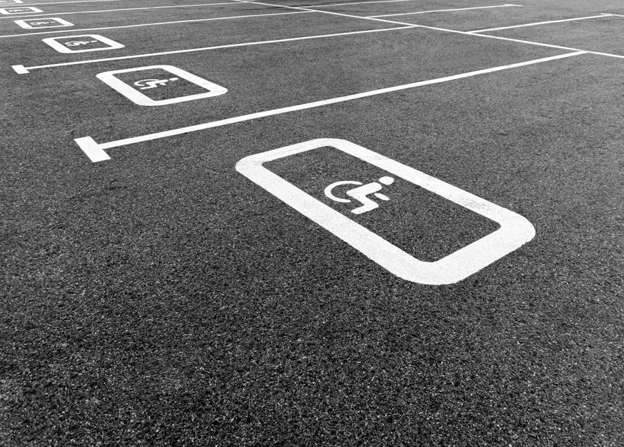 Photograph of a parking lot with clearly marked accessible parking spaces painted in white. The spaces include a prominent wheelchair symbol within rectangular outlines, indicating handicap-accessible