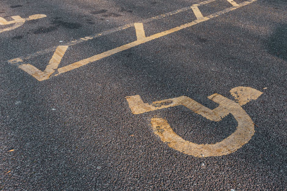 Photograph of a parking lot with clearly marked accessible parking spaces painted in white. The spaces include a prominent wheelchair symbol within rectangular outlines, indicating handicap-accessible