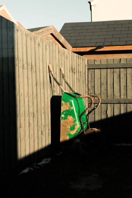 A garden scene showing a wooden fence with vertical slats casting shadows on the ground, where a green wheelbarrow with signs of rust and dirt is leaning against the fence. Behind the fence, part of a